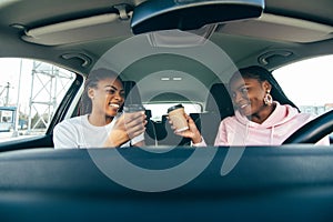 Two african woman drinkink coffee to go in the car while driving car on the road. Transportation
