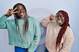 Two african woman standing over blue background doing peace symbol with fingers over face, smiling cheerful showing victory