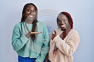 Two african woman standing over blue background showing palm hand and doing ok gesture with thumbs up, smiling happy and cheerful