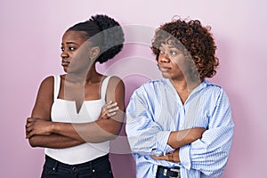 Two african women standing over pink background looking to the side with arms crossed convinced and confident