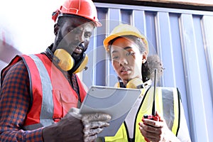 Two African workers wear safety vest and helmet at logistic shipping cargo containers yard. Happy young engineer woman uses