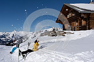 Two alpine skier and snowboarder throwing snow in air in front of Swiss chalet and blue sky