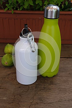 Two aluminium thermal flasks, on a wooden counter, with a vase for flowers and two green figs