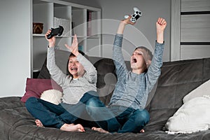 Two amazing excited teenage boys sitting on grey sofa at home, holding gaming controller, playing videogames, screaming.