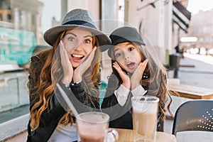 Two amazing girls in trendy hats posing with funny face expression during lunch in street restaurant in sunny day. Young