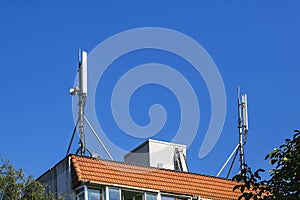 Two antennas of cellular communication on an orange tiled roof of a multi-storey residential building against blue sky