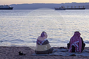 Two Arabic men wearing keffiyeh, agal and thobe are seen on the beach by the Gulf of Aqaba, photo