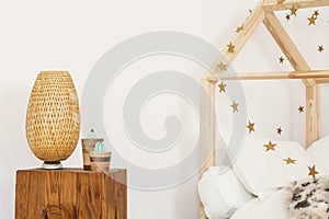 Two artificial cactuses and wicker lamp standing on wooden bedside table in white kid room interior