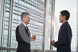 two asian business men standing and talking by the window in office