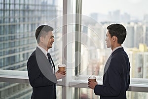 two asian business men standing and talking by the window in office