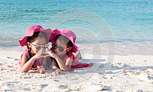Two asian child girls wearing pink hat and sunglasses playing with sand and making hand heart shape together on the beach