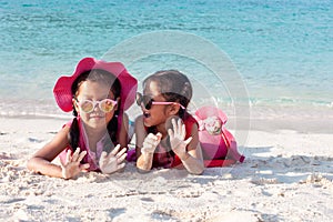 Two asian child girls wearing pink hat and sunglasses playing with sand together on the beach near the beautiful sea in