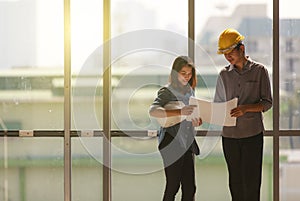 Two Asian engineer, male with yellow safety helmet and woman with white one standing and talking near high curtain wall glass