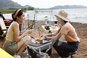 Two Asian girls having grilled barbeque enjoy camping near lake. Female enjoy fun in holiday