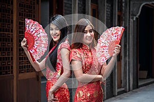 Two Asian girls wearing red cheongsams hold wooden fans in their hands.