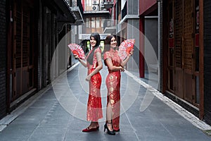 Two Asian girls wearing red cheongsams hold wooden fans in their hands.