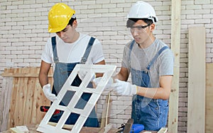 Two Asian handsome craftsmen wearing safety helmets, eyeglasses and blue overalls making DIY wooden furniture at indoor home