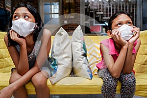 Two Asian Little Girls Wearing Medical Face Mask Getting Bored Sitting on Sofa Apart from Each Other