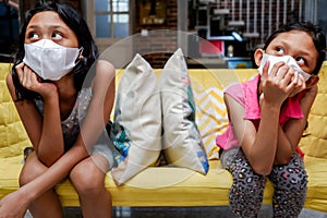 Two Asian Little Girls Wearing Medical Face Mask Getting Bored Sitting on Sofa Apart from Each Other