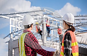 Two Asian man and woman engineer wears white safety helmet stand hold blueprint and radio while checking build in the construction