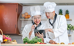 Two Asian professional couple chef wearing white uniform, hat, preparing ingredients, cooking meal in kitchen together, using