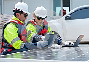 Two Asian professional technician workers sit near solar cell panel and use tablet and laptop to work in concept of green energy