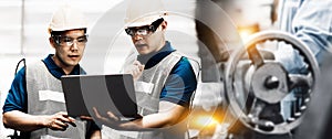 Two Asian workers talking in a metal fabrication plant wearing hardhats and protective eyewear.