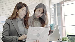 Two Asian young creative businesswomen holding a cup of coffee, working on laptop and disscuss about work while working in office.