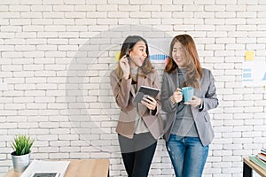 Two Asian young creative businesswomen holding a cup of coffee, working on laptop and disscuss about work while working in office