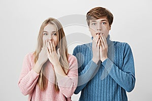 Two astonished horrified young students in stylish sweaters staring at camera in full disbelief, keeping mouths opened