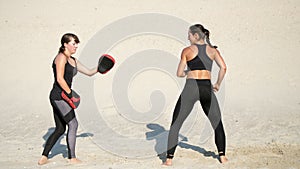 Two athletic, young women in black fitness suits are engaged in a pair, work out kicks, on a deserted beach, against a