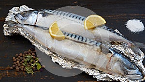 Two atlantic mackerel fishes ready for cooking decorated with spices, salt and lemon slices on the black wooden table close up
