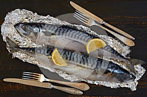 Two atlantic mackerel fishes ready for cooking decorated with spices, salt, lemon slices, knife and fork on the black wooden table