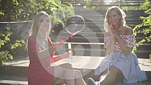 Two attractive beautiful young blond women blow bubbles in the park and are happy on sunset. Girlfriends in park on the steps