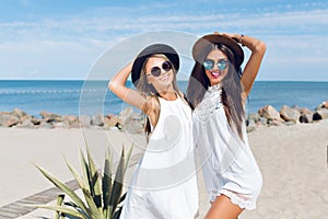 Two attractive brunette and blond girls with long hair are standing on the beach near sea. They wear hats, sunglasses