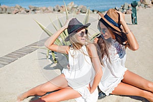 Two attractive brunette and blonde girls with long hair are sitting on the beach near sea. They wear hats, sunglasses