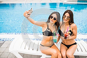 Two attractive brunette women wearing bikini posing near the swimming pool, making selfie photo. Summer time
