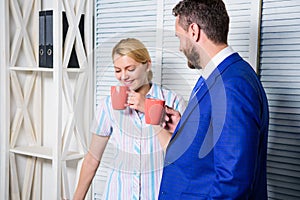 Two of attractive business people, standing next to each other, holding a cups, smiling standing at office. Job