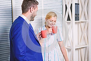 Two of attractive business people, standing next to each other, holding a cups, smiling standing at office. Job