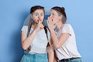 Two attractive dark haired young women posing against blue wall, lady with knot whispers to her mate shocking news, female with