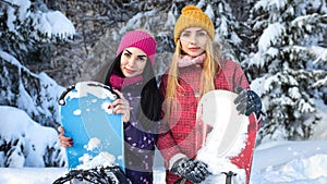Two attractive girls snowboarders are among snowy fir trees in the winter and keep snowboards hands