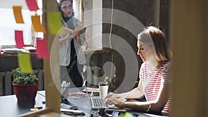 Two attractive girls are working together in modern loft office. Blonde woman is typing on laptop sitting at table while