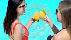 Two attractive happy women clinking beverages at summer party near swimming pool