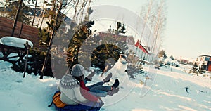 Two attractive happy young women smile sledging on snow slope towards sunny house. Winter holiday leisure slow motion.