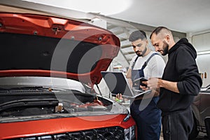 Two attractive mechanics men standing and looking at laptop with tablet and under car hood,