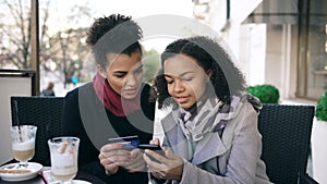 Two attractive mixed race women having online shopping with credit card and smartphone while talking and drinking coffee