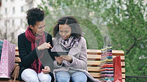 Two attractive mixed race women with shopping bags sitting on bench and using tablet computer for internet surfing