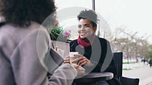 Two attractive mixed race women talking and drinking coffee in street cafe. Friends have fun after visiting mall sale