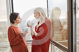 Two attractive and sensual girlfriends standing near opened window in red clothes while drinking coffee