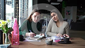 Two attractive smiling businesswomen working together while sitting at the cafe indoors